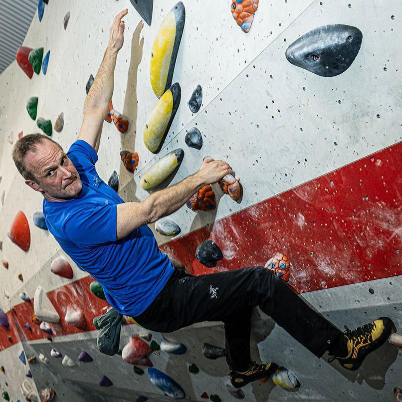 A man with brown hair wearing a blue t shirt and black tracksuit bottoms and holding onto an indoor climbing wall. He has a look of concentration on his face.