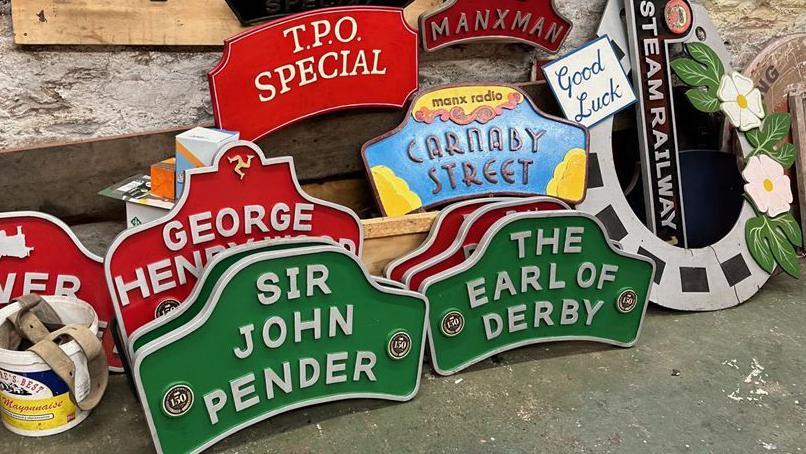 Name place of the Isle of Man Steam Railway engines stacked in the railway workshop. They are mainly green or red and feature letter including Sir John Pender and The Earl of Derby. There is also one in blue and yellow with Carnaby Street written on it, and another in the shape of a horseshoe with the words good luck on it. 