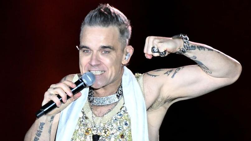 Robbie Williams sings into a microphone on stage. He smiles wearing a sparkly vest