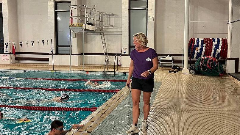 Head coach of West Norfolk Swimming club Sarah Vanderloo stands on the side of the pool looking at the swimmers inside. She is wearing a purple t shirt, black shorts and white trainers. 