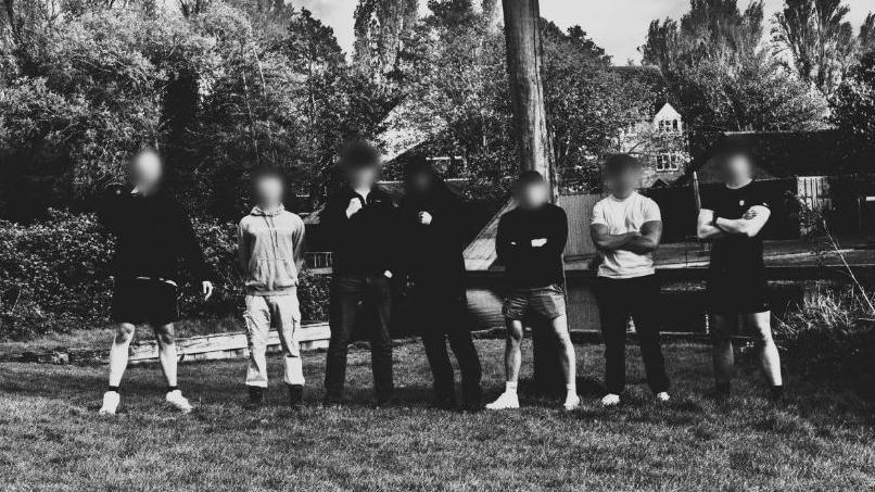 Seven members of Active Club North West pose in sportswear with their arms crossed in a rural setting for a group black and white photograph 