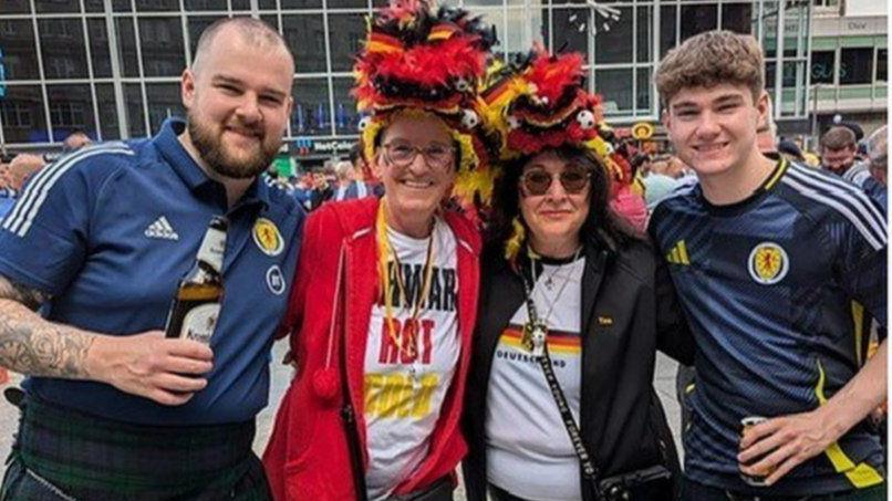 Martina Spiech, in a Germany football jersey, a red jacket and a hat in the Germany colours of red, yellow and black, poses with a similarly dressed friend, and two Scotland fans in their Scotland tops in the town square ahead of the Cologne game.