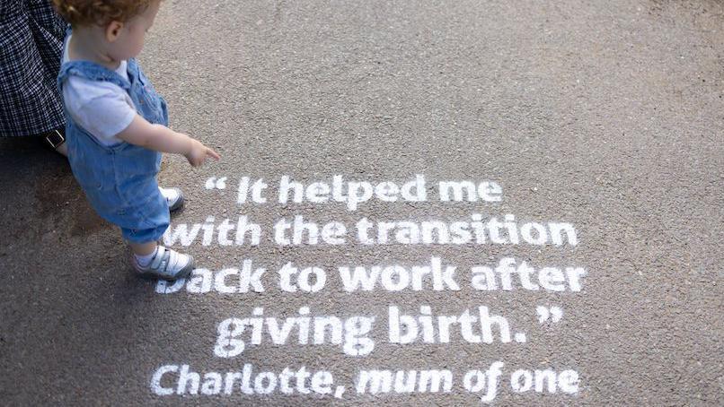 A toddler wearing denim dungarees, a white t-shirt and white velcro trainers points at words spray-painted onto a path, which reads: 'It helped me with the transition back to work after giving birth.' Charlotte, mum of one