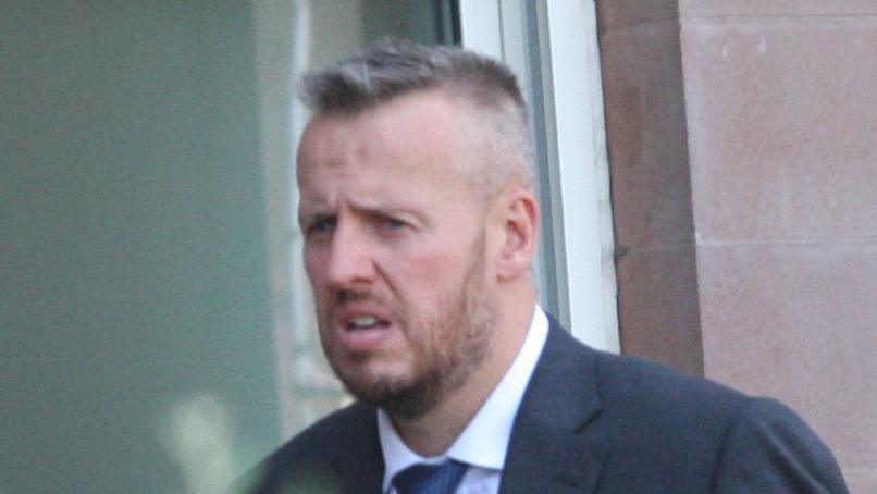 A man with short spiky hair and beard leaves court, wearing a blue suit and tie