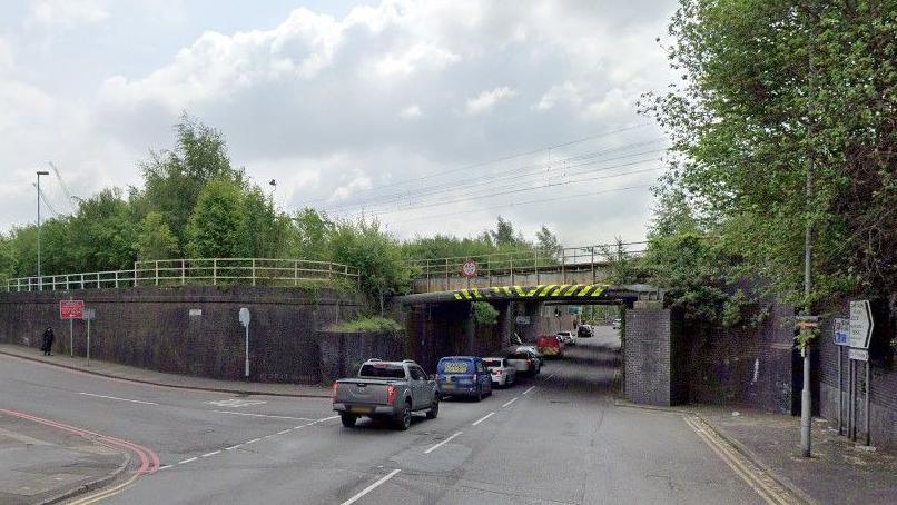 A road junction near a railway bridge with double red lines on the road on the left and double yellow lines on the road which is at right angles to it. There is queuing traffic underneath the railway bridge.