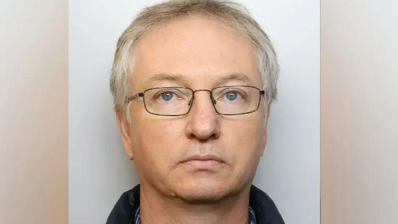 A custody picture of Chapman. He has no expression on his face, and wears dark, thin-framed glasses.