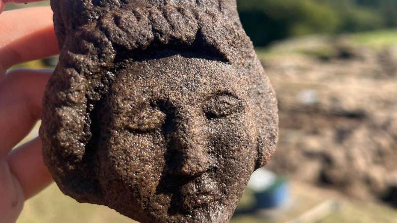 Brown stone carving of a woman's head. She has her eyes closed and has an elaborate hairstyle of plaits around her head. She is being held in a person's hand and three fingers are visible giving an indication of her size. 