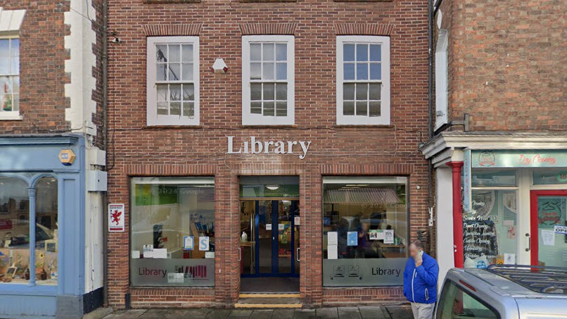 A google image of Wellington Library - a brick building in between two shops that says Library on it in white letters. There is a man in a blue jacket on his phone and a silver car to the right of the image