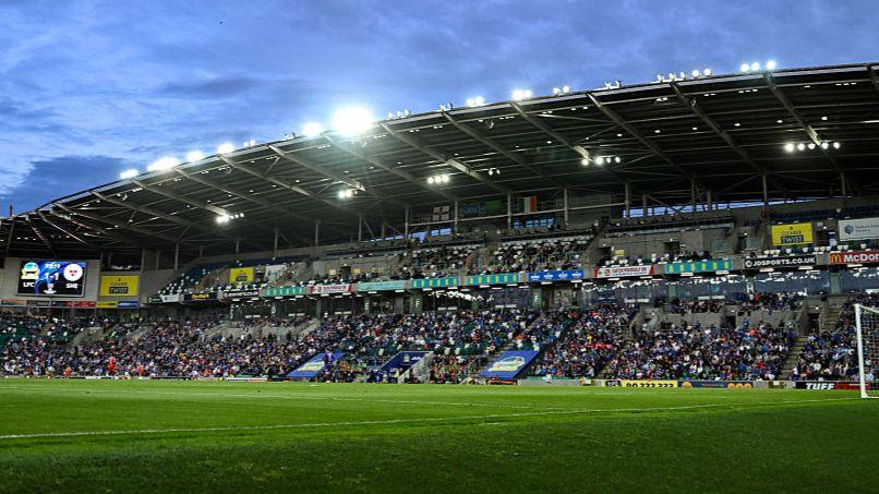 A large football stand filled with supporters. There is a grass pitch in front of the stand. The floodlights are on and the sky is blue.