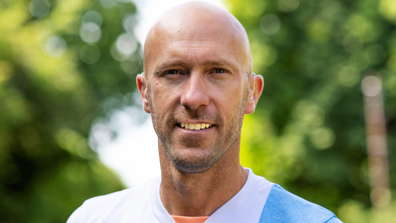 A close-up professional shot of Lewis, smiling to camera, wearing a pale blue and white sports vest and with blurred trees in the background. He is bald with some facial stubble.
