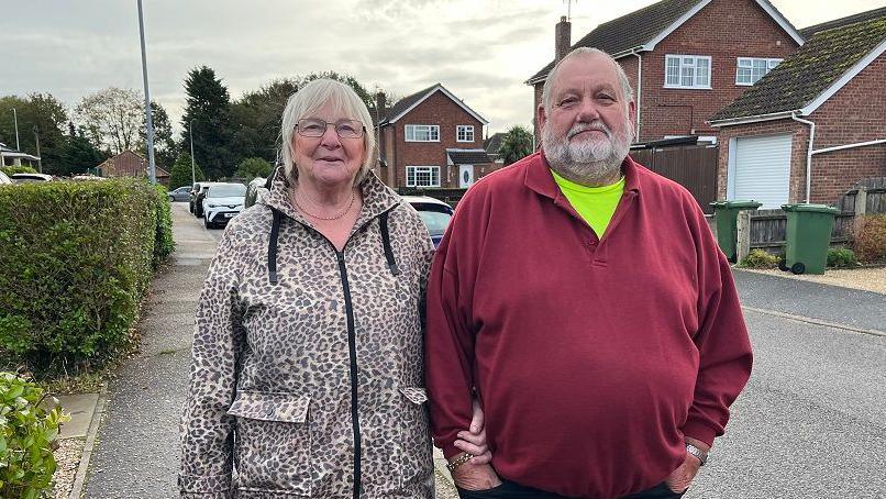 Sue and Tony Bojko stand outside Toftwood Medical Centre. Sue is wearing a leopard print coat and black leggings. Tony is in a red top with a bright yellow T-shirt under it and dark blue trousers. He has his hands in his pockets, but she has wrapped her hand under his arm and is holding his wrist. 