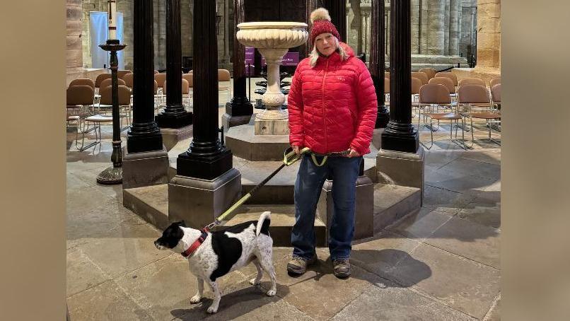 A woman in a red jacket with a red hat and a dog on a lead with a black and white fur. They are standing inside Durham Cathedral.