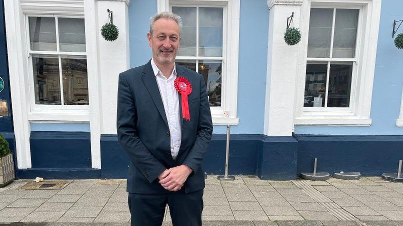 Labour Candidate for the North West Norfolk Tim Leaver stands in front of a blue building, wearing a suit and a red rosette pinned to his chest