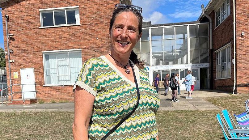 Kat Carter is wearing a yellow and green patterned top. Her sunglasses are on her head and her dark brown hair is in a ponytail. She is outside on a sunny day with the Charles Burrell building behind her.