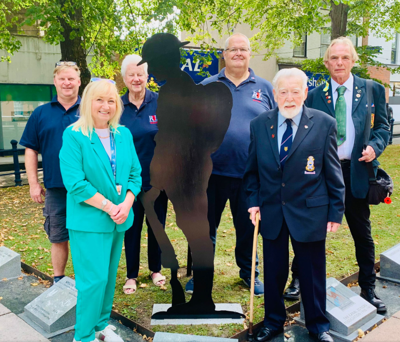 Six people stand around a metal silhouette of a soldier in the memorial gardens