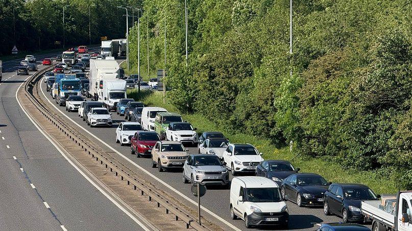 A solid queue of cars, vans and lorries heading from the top left to the bottom right of the picture. There are a few vehicles heading in the opposite direction. There is a low barrier between the carriageways. There are trees to the right.