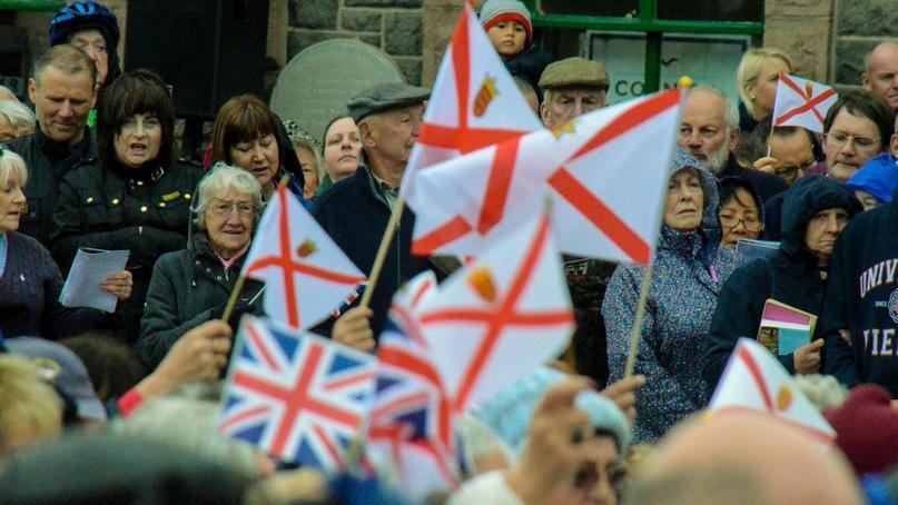 A crowd of people gathering for Liberation day celebrations. Multiple Jersey and British flags are being waved by people. As crowds look on