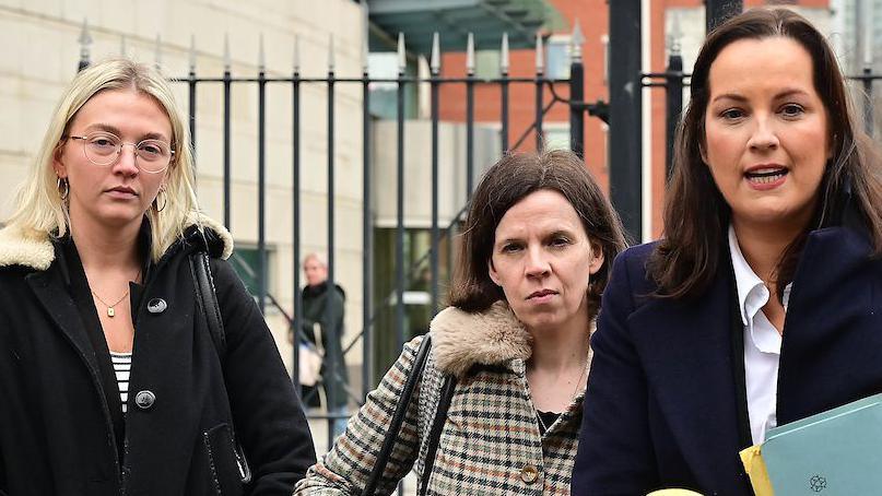 Three woman standing at microphones, one of them speaking. They are each wearing long winter coats and carrying handbags. The woman who is speaking is holding some folders in front of her. Behind them some black metal railings and behind that some buildings