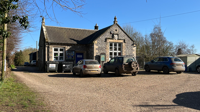 Great Dunham Primary School is a flint clad building with cars parked in front of it. It is a sunny day, with a completely blue sky above.