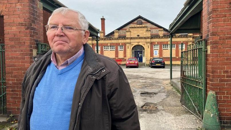 A close up picture of Jim Duffy who is stood outside a building with green gates. He is wearing a blue jumper and a dark-coloured jacket and has glasses on. 