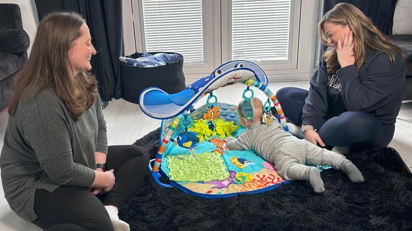 Two women sitting on the floor of a house, looking at a child on its front, on an activity mat. The woman on the right has her hand to her face, the woman on the left is smiling and looking at the child. They are both wearing dark clothing. They are all sitting on a dark rug. There are doors behind them. 