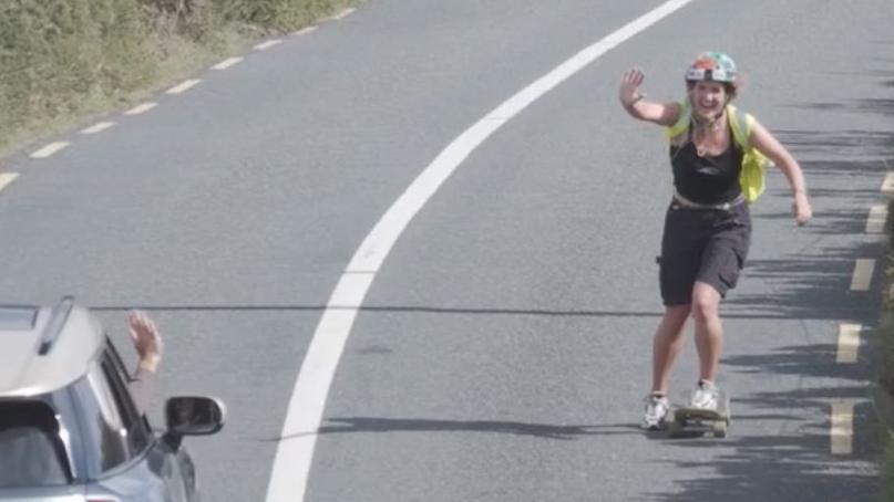 A woman is travelling along a rural road on a skateboard to the right, wearing a black vest, shorts, and a high-vis. She is also wearing a helmet, waving at an oncoming grey mini cooper on the left where the driver's hand is waving back. It appears to be a sunny day.