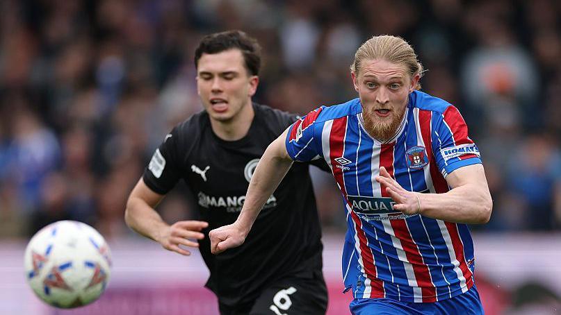 Charlie O'Connell (Boreham Wood, number six) is chasing after Luke Armstrong with his eyes closed as the Carlisle forward has his eyes on the ball in the left hand corner of the photo.