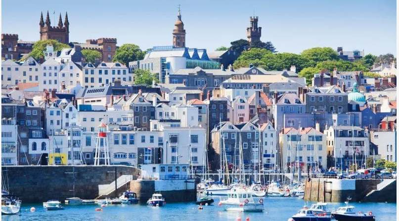 The town of St Peter Port in Guernsey. There are boats in the foreground on the water and behind it lies the town, with colourful buildings stacked on a hill.