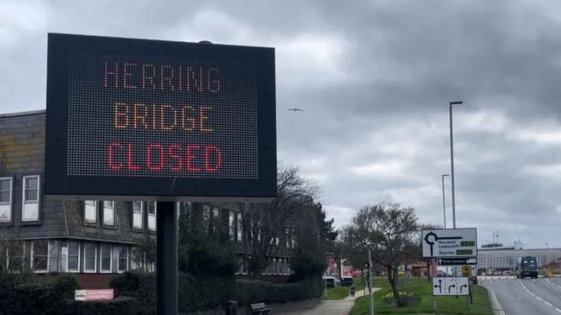 A large  sign at the side of a main road says 'Herring Bridge closed'.