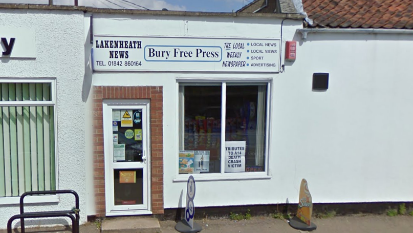 A view outside Lakenheath News. It is a white building with a single door entry and a large window next to the door.