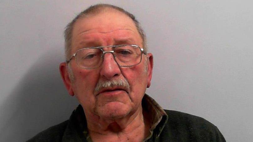 A police mugshot of a man in his early eighties. He has very short grey hair, a ruddy complexion, glasses and a short moustache.