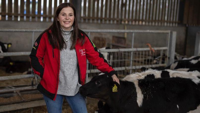 Sara Roberts mewn siwmper a jaced cynnes yn sefyll nesa i llo mewn beudy yn gwenu at y camera