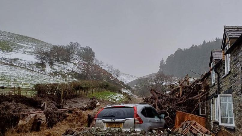 A stone house is partially obscured by a pile of fallen trees which has crashed into the outer wall. Wooden furniture can be seen lying on the floor outside the house while a silver Honda is parked outside. Hills lightly covered with snow can be seen in the background.