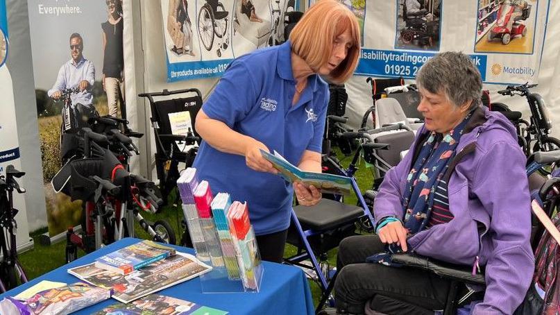 A female volunteer shows a leaflet to a female wheelchair user in a marquee containing a number of mobility walkers and scooters