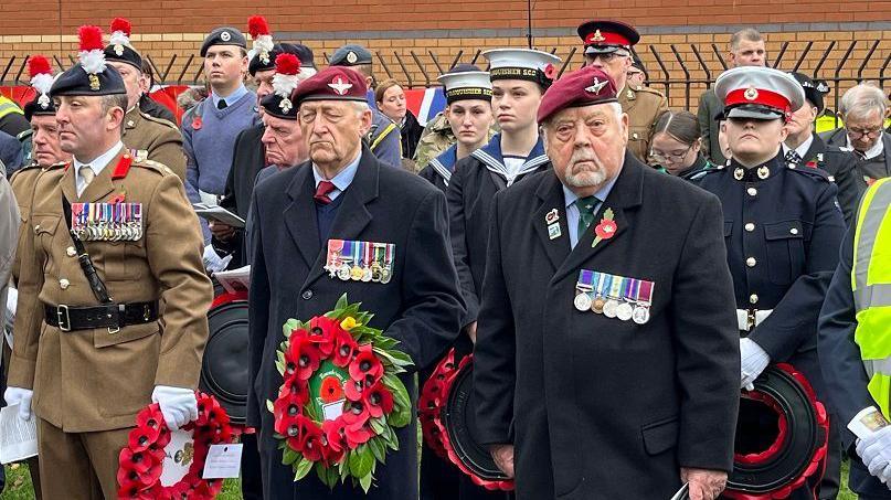 Veterans stand with poppy wreaths for the service at Bedworth. They are wearing military berets and medals. Younger personnel are also at the Armistice Day event.