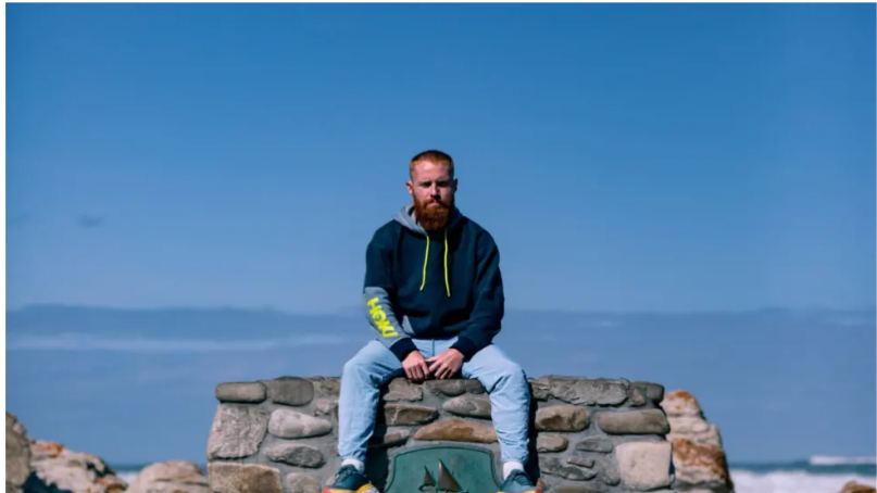 A man sat on a rock staring at the camera. He has ginger hair and a beard. He is wearing a blue hoodie and blue jeans. 