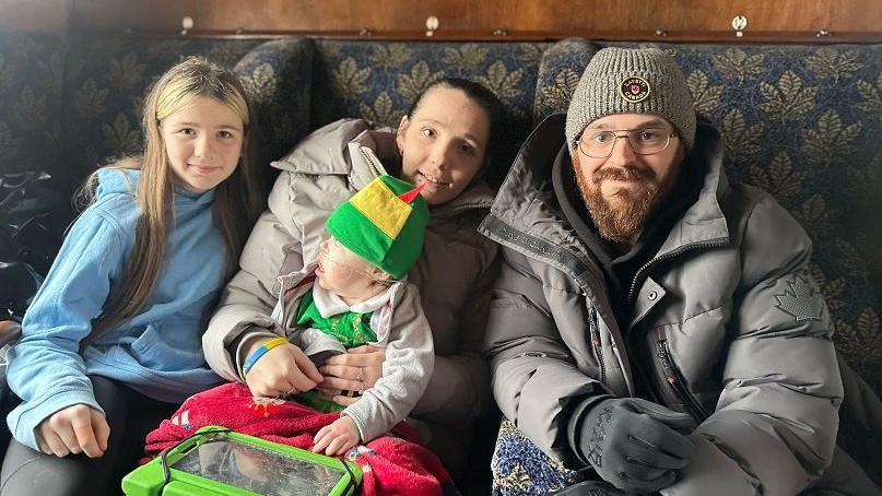 Lucy and Josh with their son Freddie and his sister are sitting on a railway seat. Freddie is wearing an elf hat and looking towards the window.