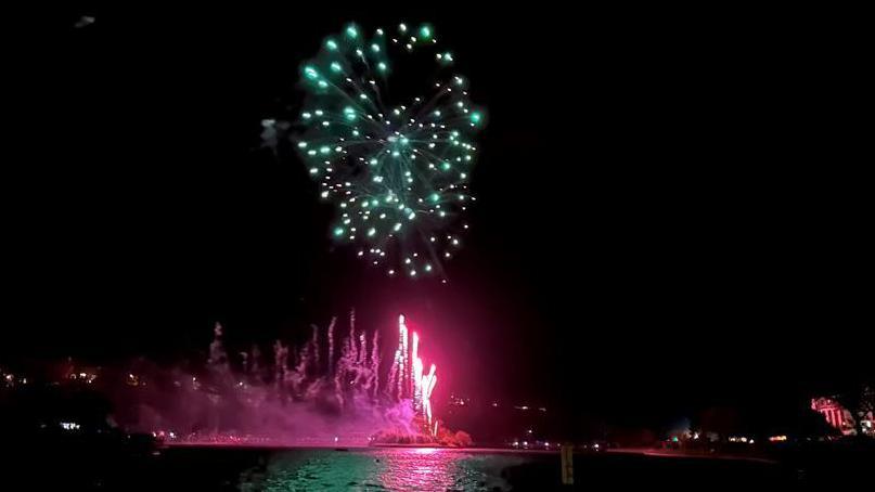 A green firework exploding over the lake at Mooragh Park in Ramsey with pink fireworks shooting up below it from the island in the middle of the lake, with the backdrop of a dark sky.