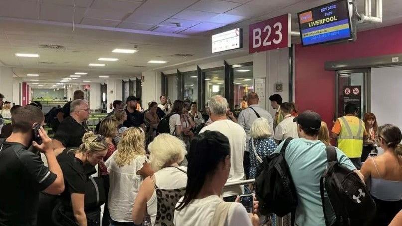 A crowd of people stood at an airport departure gate. A red sign says B23 in white writing. A screen says Liverpool in yellow writing next to the Easyjet logo of white writing on an orange background.