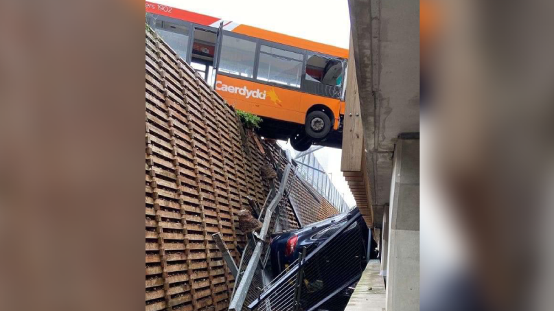 A bus is left hanging over a gap. Metal railings and a car are visible at the bottom of a chasm/gap between a carpark and a multi-storey car park. A 