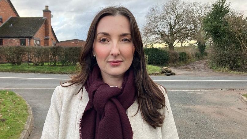 A woman with long dark hair past her shoulders wears a light white coat with a purple scarf knotted around her neck. She stands on a road leading to a main road and behind the road is a red brick building next to trees and a muddy path