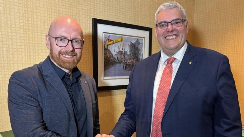 DUP leader Gavin Robinson seen shaking hands with Darryl Wilson. 
Both men are wearing suit jackets and a framed picture can be seen behind them