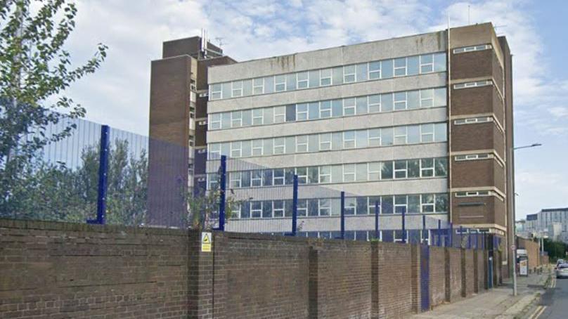 A five-storey brick building with large windows behind a brick wall with blue metal fencing.