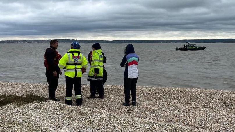 Two people in high-vis coats with two others standing next to the shoreline with a boat in the river