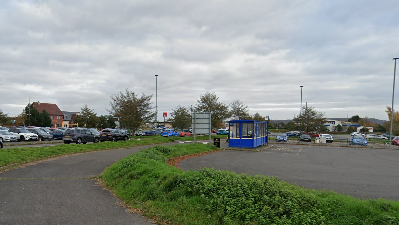 A large car park filled with cars. There is a curved road area for buses, and a blue bus shelter.