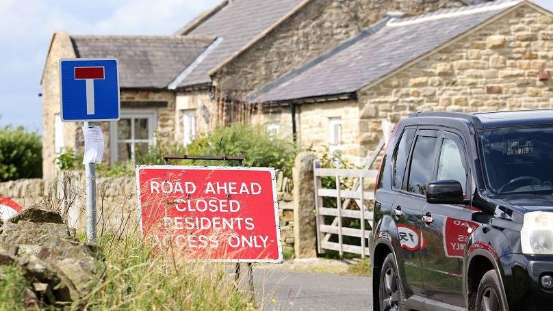 A village lane with a one-storey stone cottage, gate and partial view of a car, a cul-de-sac sign and another stating that the road ahead is closed with residents access only.