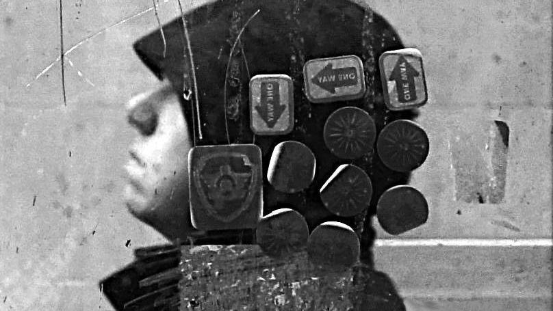 A hooded figure is photographed through a cracked and stained glass. The image is in black and white, and several stickers saying "one way" and containing symbols are on the glass