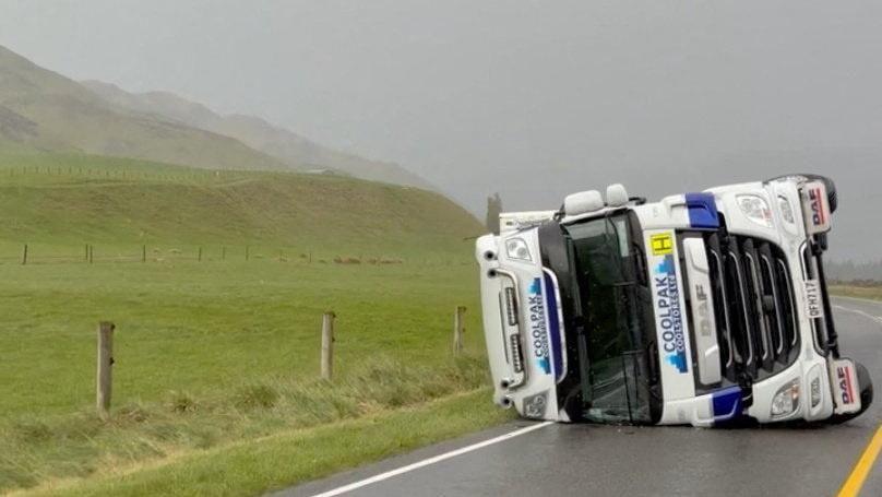 An overturned white truck lying on its side on a grey road, next to a grassy meadow that is fenced off.