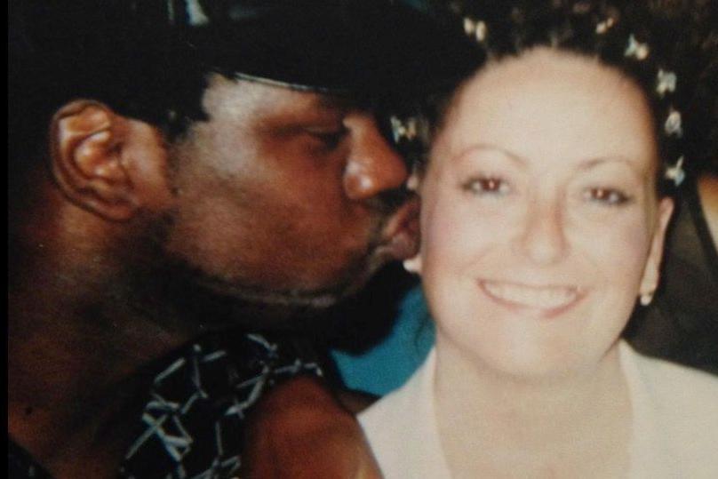Winbourne and Carole Charles, seemingly at a party or event. Mr Charles is kissing the side of Mrs Charles's face as she smiles at the camera. She has decoration in her hair, pearl earrings and a white top. He is wearing a black shirt with a white pattern on it.