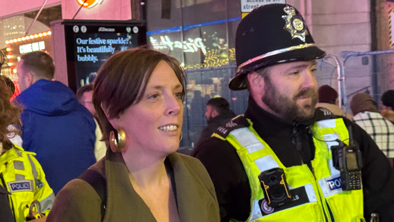 A woman with brown hair and large gold ear rings in a green coat stood in a town centre next to a bearded police officer with black uniform and a yellow reflective vest